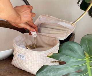 Hand placing a beauty item into an Astala Luna Vanity Case beside a sink and green plants.